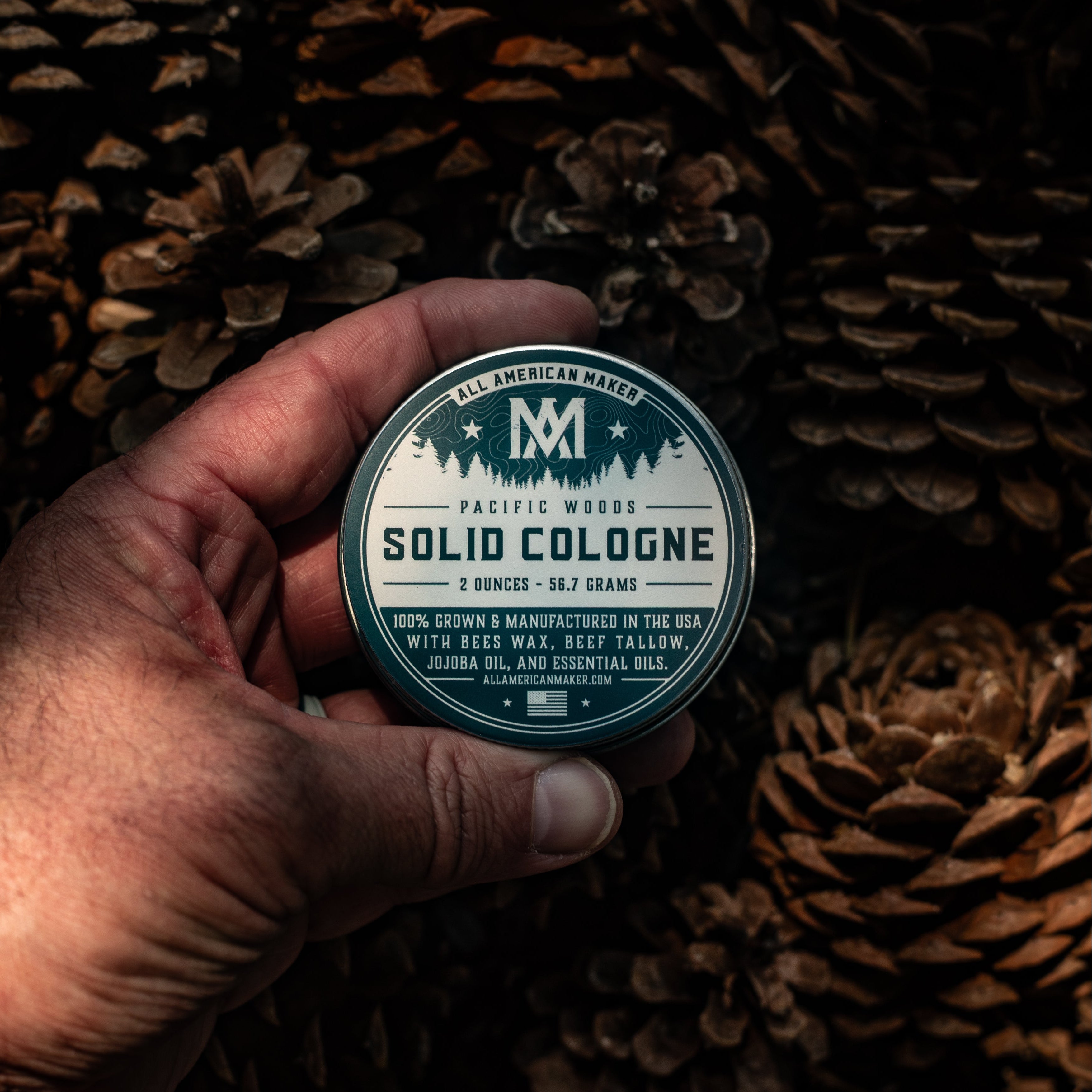 A hand holds a round tin of Pacific Woods Solid Cologne against a dark background of pinecones. The tin has a white label with navy blue text and graphics.
